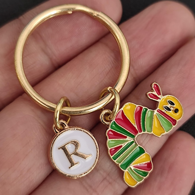 Puede incluir: Llavero dorado con un dije circular blanco con la letra "R" y un dije de oruga colorido. La oruga es principalmente roja, amarilla y verde. Un accesorio decorativo.