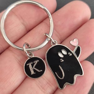 May include: A silver keychain with a round charm featuring the letter "K" and a black ghost charm holding a pink heart. The ghost has white eyes and a curved mouth. The keychain is held in a hand.