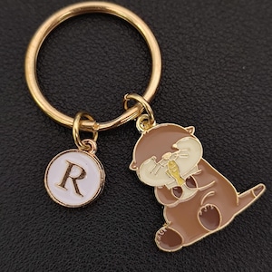 May include: Gold-coloured keyring with a round charm featuring the letter "R" in white, and a cartoon otter charm in brown, tan, and yellow. The otter is holding a fish. The keyring is on a dark surface.
