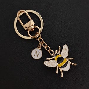 May include: Gold-tone keychain featuring a bee charm with black, yellow, and white enamel detailing. Includes a small white circular charm with the letter "N". The keychain has a lobster clasp and a key ring.
