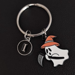 Könnte beinhalten: Ein Halloween-Schlüsselanhänger mit einem weißen Geist, der einen orangefarbenen Hut trägt und eine Sense hält. Der Schlüsselanhänger enthält auch einen kreisförmigen Anhänger mit dem Buchstaben "I" auf schwarzem Hintergrund, alles an einem silbernen Schlüsselring befestigt.