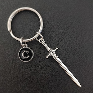 Könnte beinhalten: Silberfarbener Schlüsselanhänger mit einem Miniatur-Schwertanhänger und einem runden Charm mit dem Buchstaben "C" in schwarzem Emaille. Der Schlüsselanhänger hat einen großen Ring für Schlüssel und einen kleinen Ring zur Befestigung der Charms.