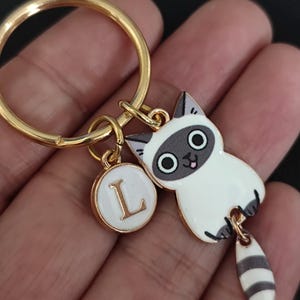 May include: Gold key chain with a white and gray cat charm with a black and white striped tail. The cat has a pink tongue sticking out and large black eyes. The keychain also has a small round charm with the letter L in gold on a white background.