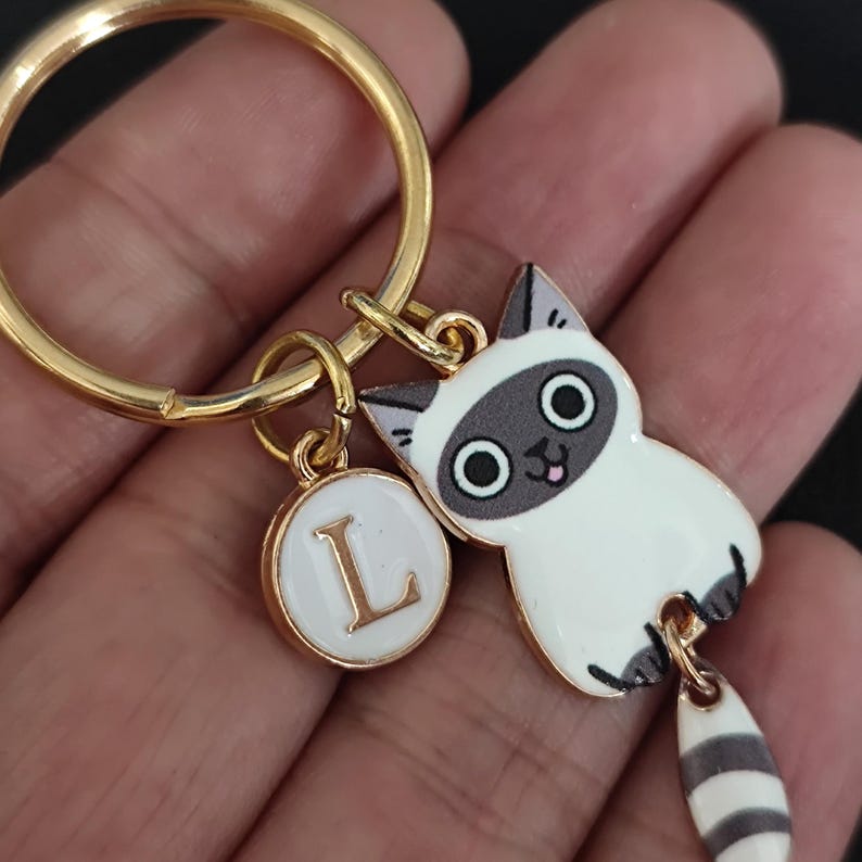May include: Gold key chain with a white and gray cat charm with a black and white striped tail. The cat has a pink tongue sticking out and large black eyes. The keychain also has a small round charm with the letter L in gold on a white background.