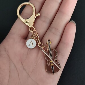 May include: A gold-colored keychain with a cello charm and a white circular charm with the letter "A". The cello charm is brown with a yellow accent and a white bow. The keychain has a lobster clasp.