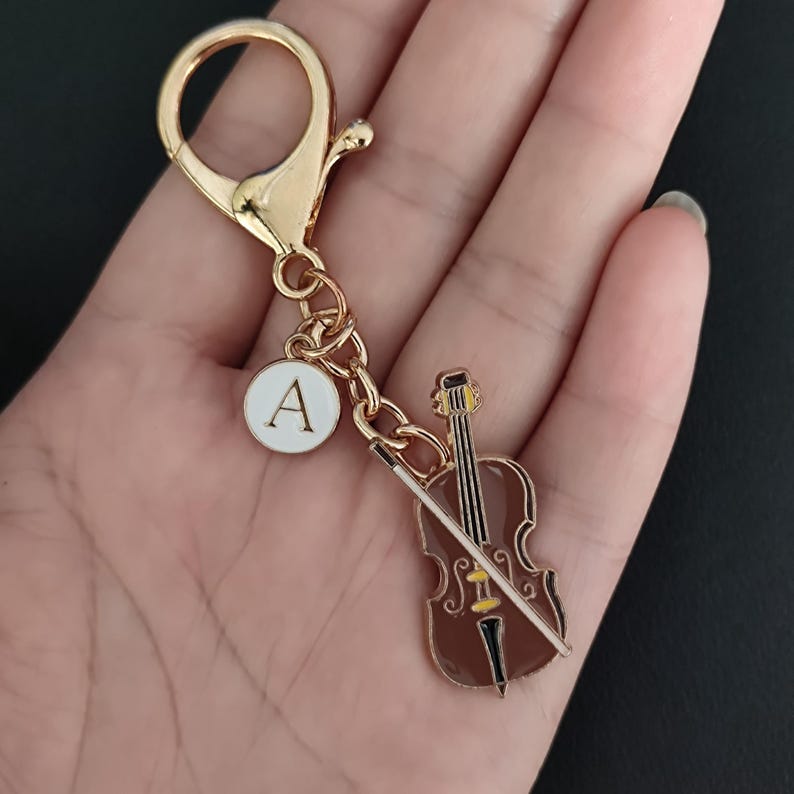 May include: A gold-colored keychain with a cello charm and a white circular charm with the letter "A". The cello charm is brown with a yellow accent and a white bow. The keychain has a lobster clasp.