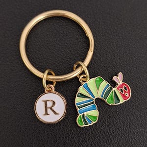 Puede incluir: Llavero de metal dorado con dos colgantes. Un colgante es un c&iacute;rculo blanco con la letra "R" en el centro. El otro colgante es una oruga colorida con detalles verdes, azules y rojos. El llavero est&aacute; sobre una superficie negra.