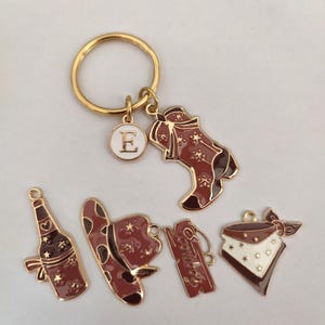 Könnte beinhalten: Ein goldener Schlüsselanhänger mit verschiedenen Anhängern, darunter ein Cowboy-Stiefel, ein Cowboyhut, eine Weinflasche, ein Bandana und ein Anhänger mit dem Buchstaben E. Die Anhänger sind alle braun und goldfarben mit weißen Akzenten.