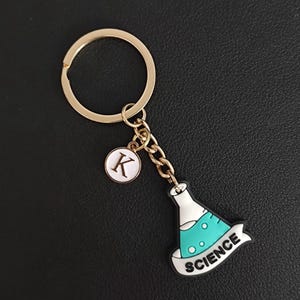 Op de afbeelding: Goudkleurige sleutelhanger met een wetenschapsthema bedel. De bedel heeft een turquoise en witte fles met het woord "SCIENCE" op een banner. Inclusief een kleine ronde bedel met de letter "K".