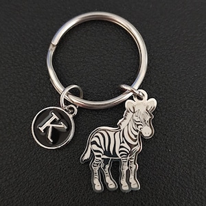 Pode incluir: Um chaveiro prateado com um charme de zebra preto e branco e um charme prateado com a letra K.