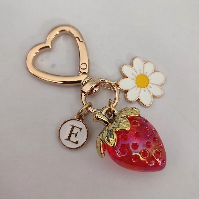 May include: Gold heart-shaped keychain with a red and gold strawberry charm, a white daisy charm, and a white circle charm with the letter 'E' in gold.
