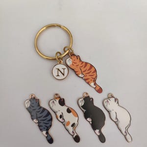 May include: A gold-colored keychain with a round charm featuring the letter "N" and a tabby cat charm. Below are four additional cat charms in various colors: gray, calico, black, and white. All charms have a gold-colored border.
