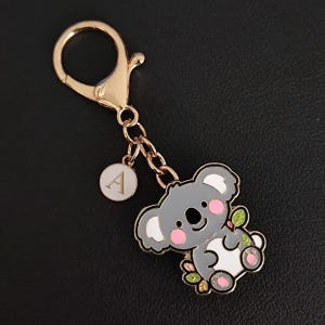 May include: A gold-colored keychain featuring a cartoon koala bear with gray fur, pink cheeks, and white accents. The koala holds green leaves and is attached to a chain and a clasp. A small, round charm with the letter "A" hangs from the chain.