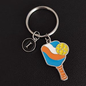 Könnte beinhalten: Ein silberner Schlüsselanhänger mit einem Pickleball-Schläger-Anhänger in Blau, Orange und Weiß sowie einem gelben Ball. Enthält einen schwarzen und silbernen Initialen-Anhänger "I". Der Schlüsselanhänger befindet sich auf schwarzem Hintergrund.