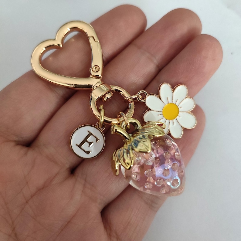 May include: Gold-tone keychain with a heart-shaped ring, a charm with the letter E, a pink and white strawberry charm, and a white daisy charm with a yellow center.