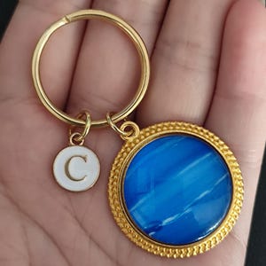 Op de afbeelding: Goudkleurige sleutelhanger met een ronde blauw-witte hanger en een witte bedel met de letter "C". De blauwe hanger heeft een getextureerd uiterlijk en is omgeven door een goudkleurige, kralenrand. De sleutelring is ook goudkleurig.