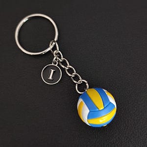 May include: Volleyball keychain with a blue, yellow, and white ball design. The keychain includes a silver ring, chain, and a black charm with the letter "I". The keychain is a sports-themed accessory.