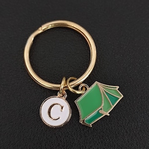 Könnte beinhalten: Ein goldfarbener Metallschlüsselanhänger mit einem runden Ring und zwei Anhängern. Ein Anhänger ist eine weiße runde Scheibe mit einem goldfarbenen Buchstaben C in der Mitte. Der zweite Anhänger ist eine grüne Zeltform mit goldfarbenen Metallrändern.