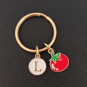 May include: Gold key ring with a white circle charm featuring the letter L and a red tomato charm with green leaves.