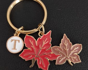 Puede incluir: Un llavero dorado con un anillo redondo y tres colgantes. Los colgantes incluyen un círculo blanco con la letra "T", una hoja de arce roja y una hoja de arce marrón. El llavero está sobre una superficie negra.