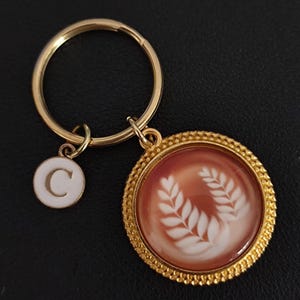 Könnte beinhalten: Goldfarbener Schlüsselanhänger mit einem runden Anhänger mit weißem Lorbeerkranz-Design auf rotbraunem Hintergrund. Ein kleinerer weißer Charm mit dem Buchstaben "C" ist ebenfalls vorhanden. Der Schlüsselanhänger liegt auf einer schwarzen Oberfläche.