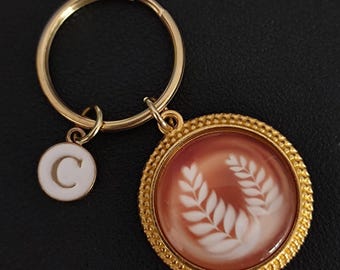 May include: Gold-toned keychain with a round pendant featuring a white laurel wreath design on a reddish-brown background. A smaller white charm with the letter "C" hangs nearby. The keychain is on a black surface.
