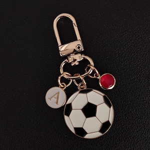 May include: Gold-tone keychain featuring a soccer ball charm with black and white panels. Includes a white circular charm with the letter "A" and a red gem. The keychain has a lobster clasp for easy attachment.