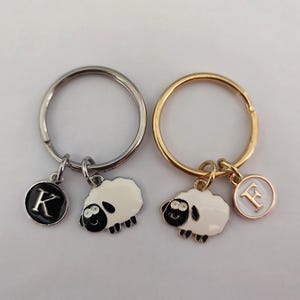 May include: Two keyrings with a sheep charm and a letter charm. The keyring on the left has a silver ring and a black letter K charm. The keyring on the right has a gold ring and a gold letter F charm. Both charms are white with black eyes and a black nose.