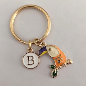 May include: Gold keyring with a toucan charm and a letter 'B' charm. The toucan is orange, yellow, white, and purple. The toucan is perched on a branch with green leaves.