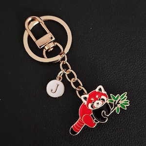May include: Gold-tone keychain featuring a red panda design with black and white accents, holding a green leafy branch. Includes a white charm with the letter "J". The keychain has a lobster clasp and a key ring.