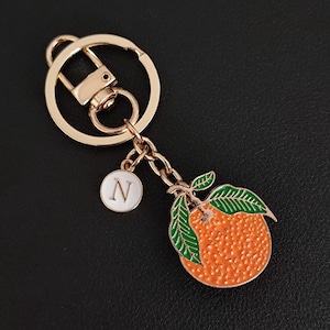 May include: Gold-tone keychain featuring an orange fruit charm with green leaves and a white enamel charm with the letter "N". The keychain has a lobster clasp and a key ring.