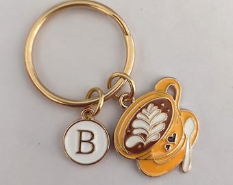 May include: Gold keychain with a coffee cup charm and a letter B charm. The coffee cup is yellow with brown coffee and white foam. The letter B is white on a gold circle.
