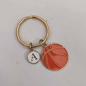 Könnte beinhalten: Goldener Schlüsselanhänger mit einem Basketball-Anhänger und einem kleinen Anhänger mit dem Buchstaben A. Der Basketball-Anhänger ist orange mit schwarzen Linien.