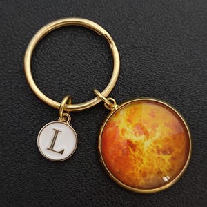 May include: A gold-coloured keyring with two charms. One charm is a white circle with the letter "L" in black. The other charm is a round pendant with an orange and yellow planet design.