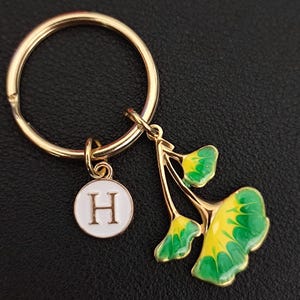 May include: Gold-tone keychain with a round charm featuring the letter "H" and a ginkgo leaf pendant. The ginkgo leaf is detailed with green and yellow enamel. The keychain is a stylish accessory.