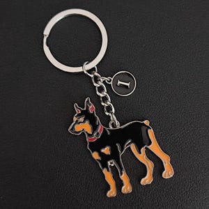 May include: A Doberman Pinscher keyring in black and orange, with a silver ring and chain. The dog has a red collar and a small black charm with the letter "I". The keyring is on a black surface.
