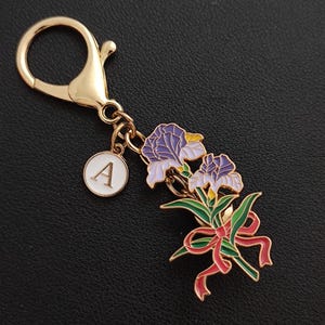 May include: Gold-coloured keyring with a floral design featuring purple irises, green leaves, and a red ribbon. Includes a white circular charm with the letter "A". The keyring has a lobster clasp for easy attachment.