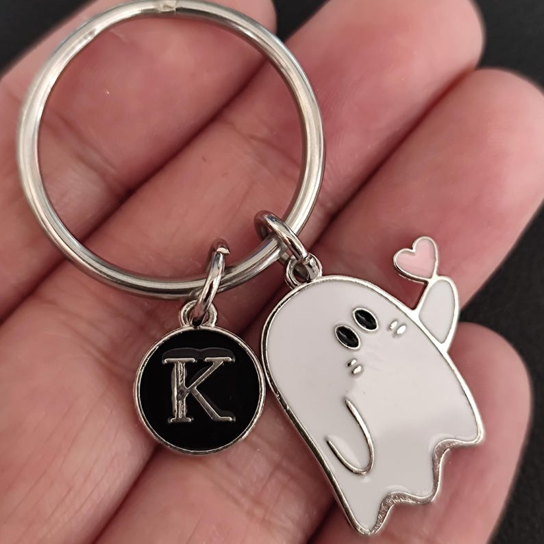 May include: Silver keychain with a white ghost charm holding a pink heart. Includes a black circular charm with the letter "K" in silver. The keychain is a cute accessory.
