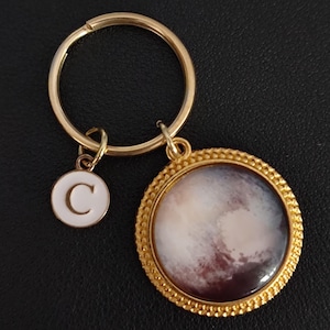 May include: Gold-coloured keyring with a round charm depicting a planetary image and a smaller charm with the letter "C". The planetary charm has a textured, multi-coloured design. The keyring and charms are gold.
