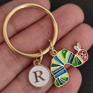 Puede incluir: Llavero dorado con un dije redondo con la letra "R" y un dije de oruga colorida. La oruga es verde, azul y roja con ojos negros. Accesorio decorativo.