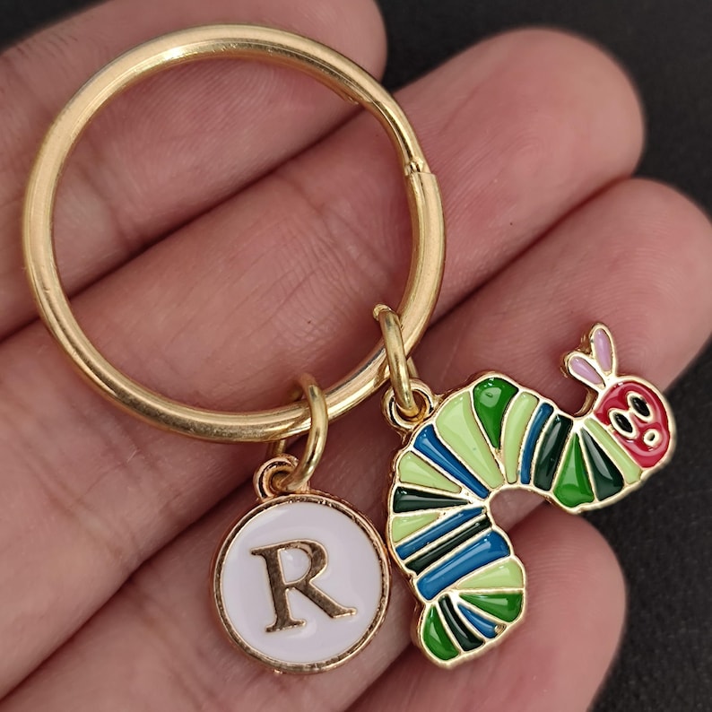Puede incluir: Llavero dorado con un dije redondo con la letra "R" y un dije de oruga colorida. La oruga es verde, azul y roja con ojos negros. Accesorio decorativo.