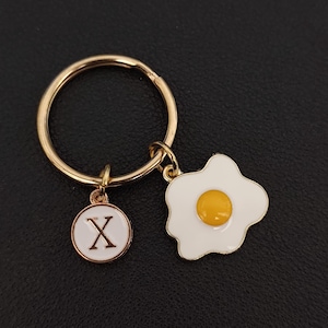 Könnte beinhalten: Goldfarbener Schlüsselanhänger mit zwei Anhängern: ein Spiegelei und ein runder Anhänger mit dem Buchstaben "X". Der Eieranhänger hat einen weißen Hintergrund mit einem gelben Eigelb. Der Buchstabenanhänger hat einen weißen Hintergrund und einen schwarzen Buchstaben.