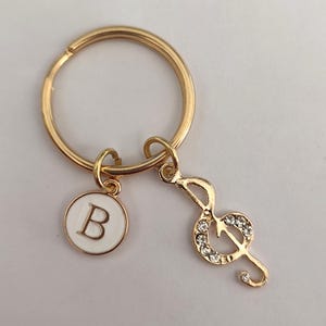 Può includere: Portachiavi dorato con un ciondolo in smalto bianco con la lettera 'B' e un ciondolo a forma di chiave di violino dorato con strass trasparenti.