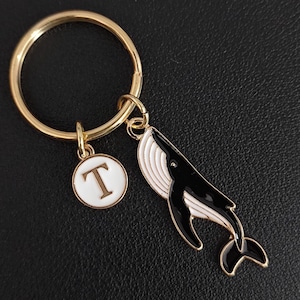 Op de afbeelding: Een goudkleurige sleutelhanger met een walvisbedel en een ronde bedel met de letter "T". De walvis is zwart en wit met goudkleurige accenten. De sleutelring is ook goudkleurig.
