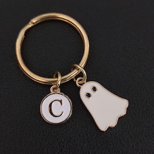 May include: Gold-tone keychain with a white ghost charm and a white circular charm with the letter "C". The ghost charm has black eyes. The keychain is on a black surface.