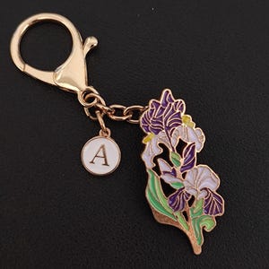 May include: Gold-coloured keychain featuring a floral iris design in purple, white, and green enamel. Includes a small white charm with the letter "A". The keychain has a lobster clasp for easy attachment.