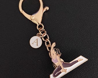 May include: A gold-colored keychain with a charm featuring a yoga pose. The charm has a white background with a woman in a purple sports bra and leggings. A white circular charm with the letter "A" hangs from the chain.
