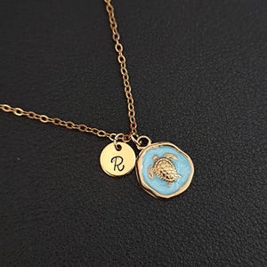 Könnte beinhalten: Goldfarbene Halskette mit einem Schildkrötenanhänger vor hellblauem Emaille-Hintergrund und einem kleinen runden Anhänger mit dem Buchstaben "R". Die Kette hat eine zarte Kette und einen Karabinerverschluss.