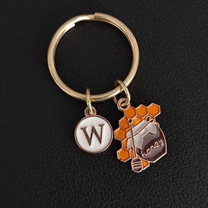May include: Gold-tone keychain with two charms. One charm is a white circle with a capital "W" in brown. The other charm is a honey pot with a honeycomb design and the word "honey".