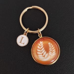 Può includere: Portachiavi dorato con due ciondoli. Un ciondolo è un cerchio bianco con la lettera "L" in oro. L'altro è un ciondolo rotondo con uno sfondo color caffè e un disegno di foglie bianche. L'anello del portachiavi è anch'esso dorato.